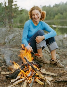 Young woman sitting at camp fire, holding fried sausage
